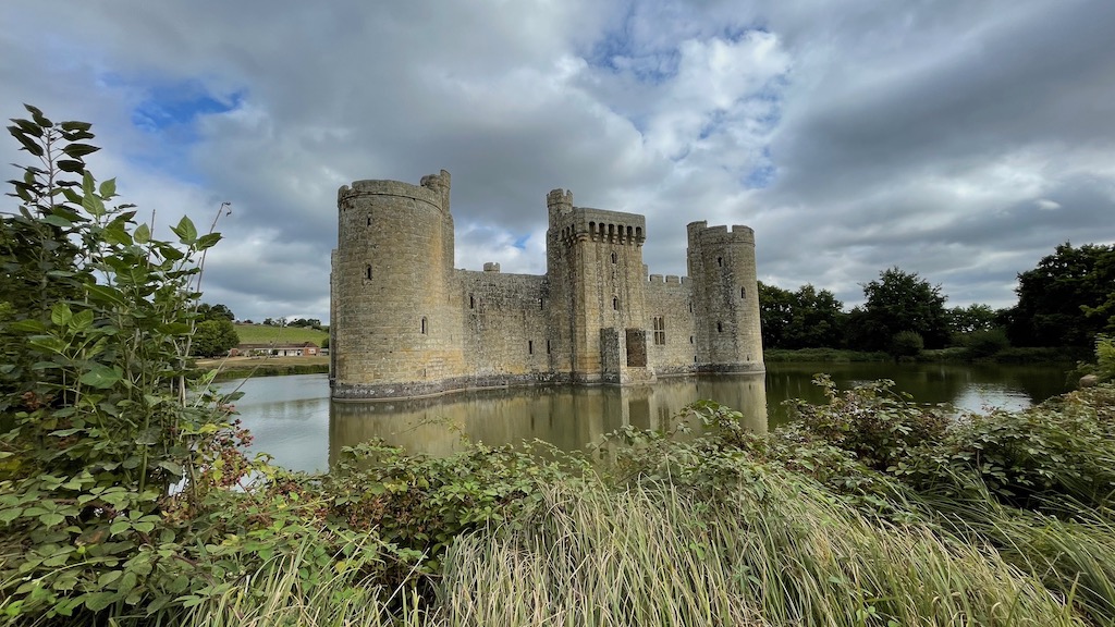 A castle standing in the middle of a moat seen through some greenery. The sky is blue with some ominous clouds