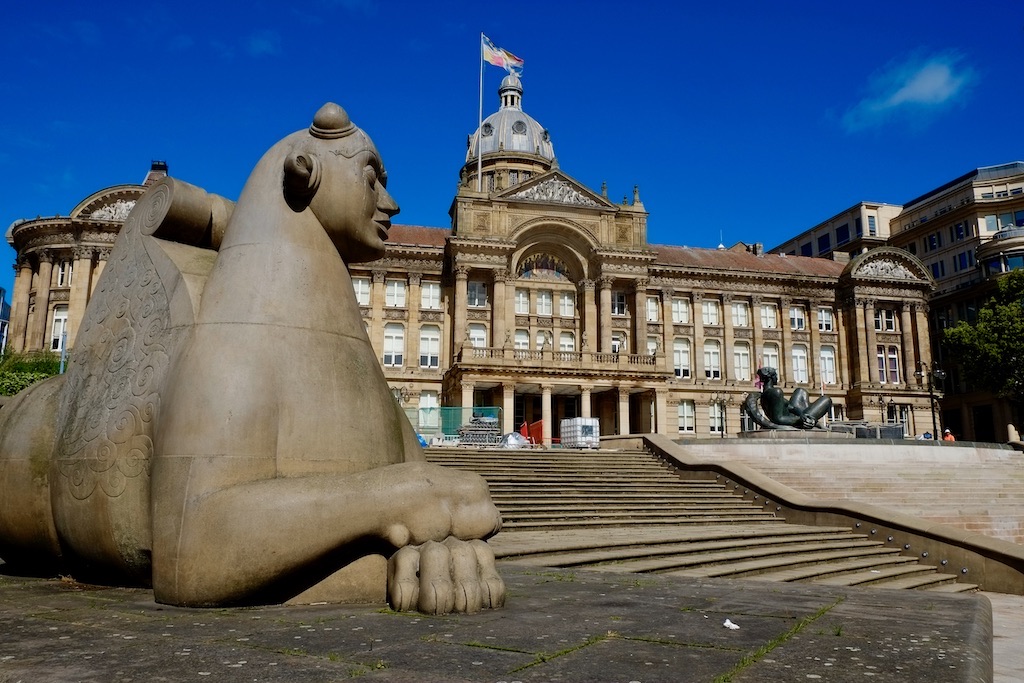 A bulbous lion statue reclines in the foreground, with a large victorian building behind