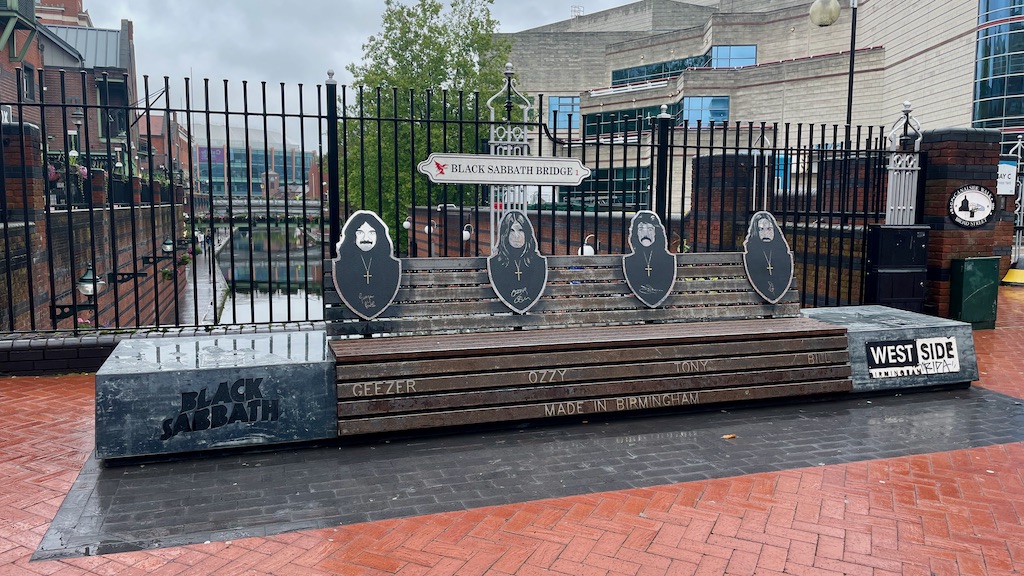 A large bench sits on a bridge in rainy Birmingham. On the bench are four silhouettes of the members of Black Sabbath. 