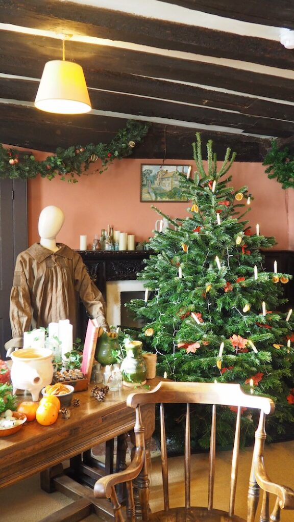 A christmas tree in a kitchen with a mannequin next to it. The wall behind is a dark peach.