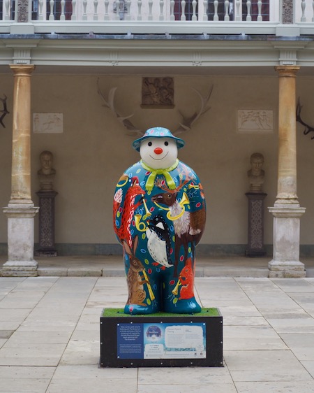 A snowman in a courtyard. The antlers on the wall behind are lined up perfectly with his head 