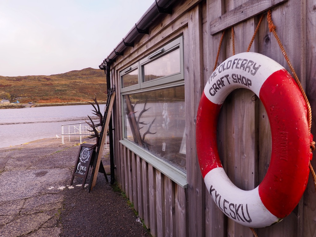 A wooden shack to the right of the image with a lifebuoy hanging from the side and some antlers leaning against it. The background view is a loch