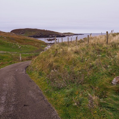 A road between two grass banks. It sneaks around a corner down a hill and in the distance you can see the sea.
