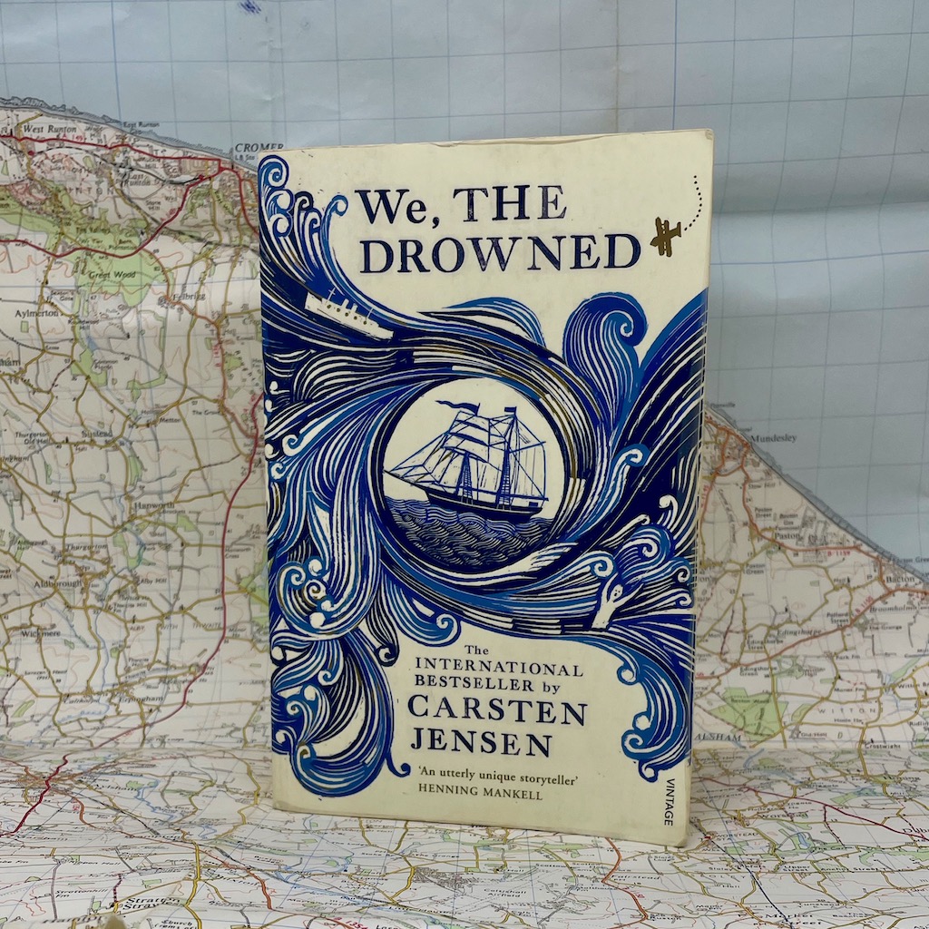 A book called We the drowned sits in front of a map. The book has blue waves circling an old sailing ship against a cream background.