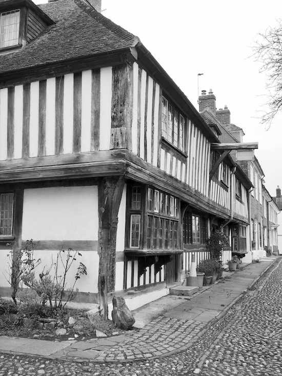 A black and white photograph of a timber framed house on a street corner. The street is cobbled