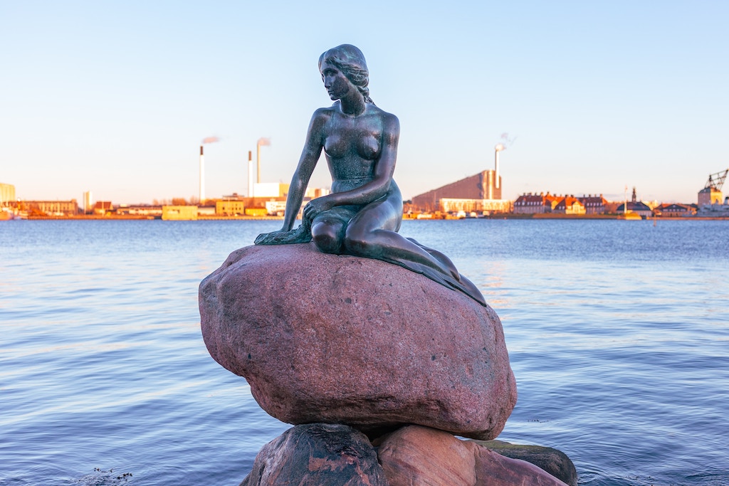 A statue of the little mermaid from the fairytales sits on a rock in a harbour.