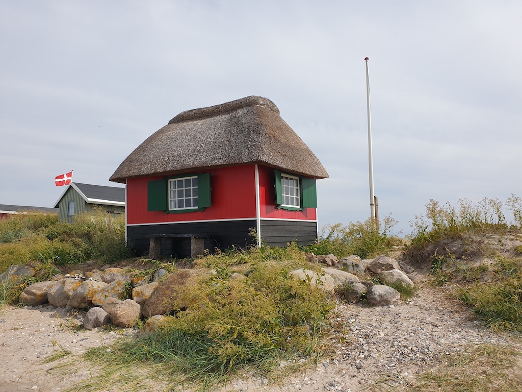 A red hut sits on the crest of a hill. It has a thatched roof and green shutters.