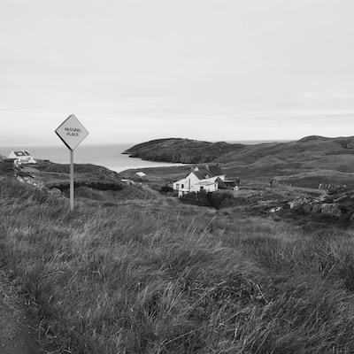A passing place sign to indicate a narrow road. In the distance is a white house and a beach